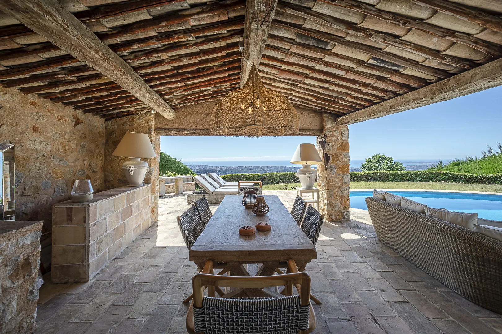 Panorama : terrasse couverte sous une charpente traditionnelle, table de repas et piscine face à la mer